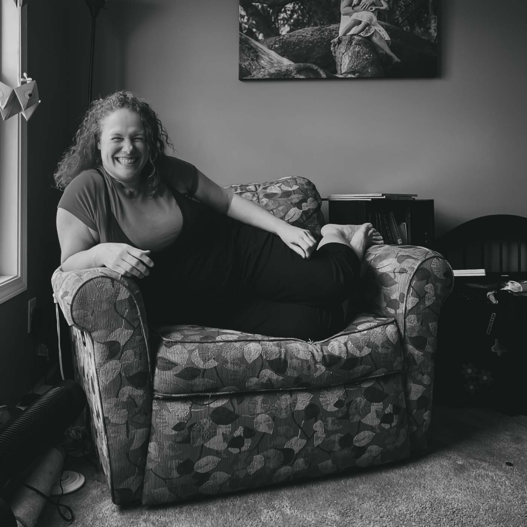 black and white portrait of Mell Bell lounging across a large recliner chair with one arm on the arm rest and her two feet crossed at the ankles on the other arm rest. She is laughing and smiling so big that her nose is crinkled and her eyes are closed.