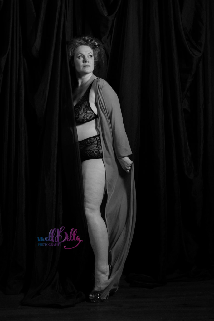 black and white full length portrait of the photographer turned to the side wearing a lace bra and panty set and a long robe. Her body is half hidden behind a sheer black curtain, and she looks off to the right side of the image. Her expression is pensive and a little unsure.