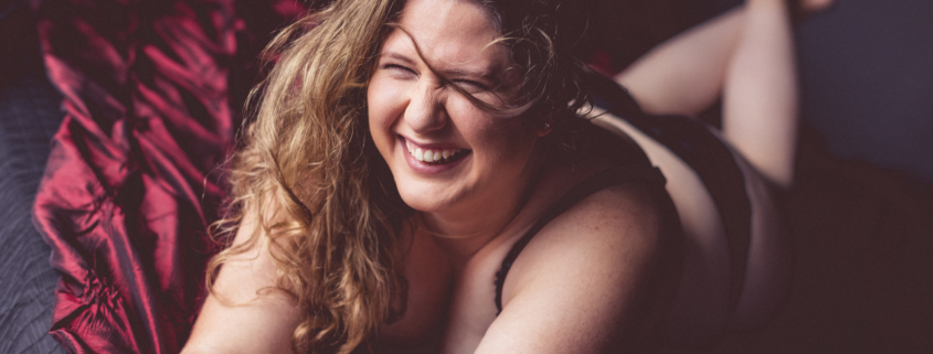 Smiling woman laying on stomach with her long light brown hair tousled to one side. She is laying on a burgundy textured cloth and is wearing a black outfit.