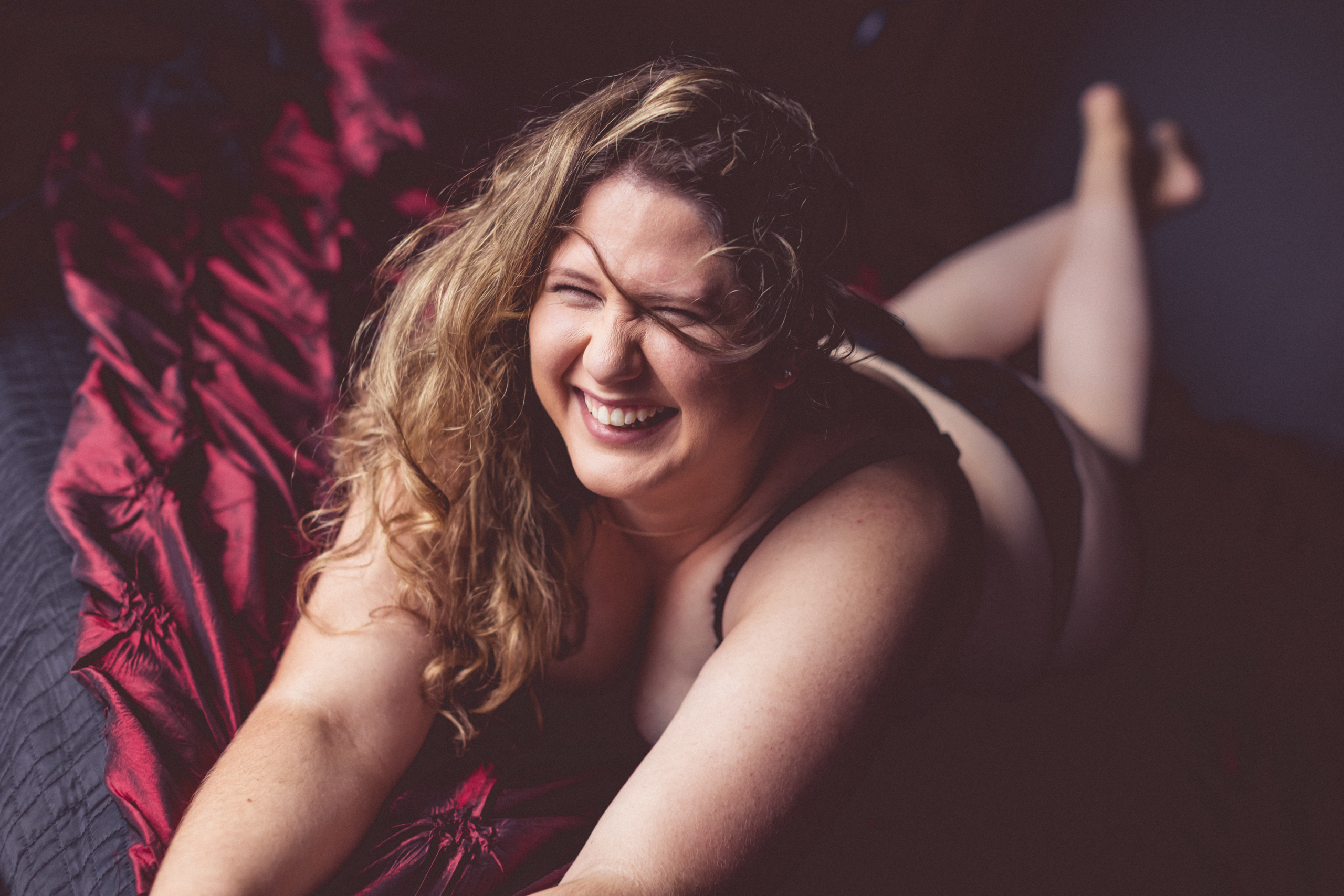 Smiling woman laying on stomach with her long light brown hair tousled to one side. She is laying on a burgundy textured cloth and is wearing a black outfit.