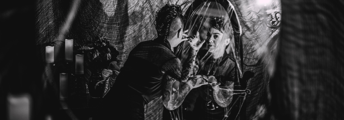 black and white image of a halloween styled room with spider webs, candles, a woman looking in a mirror, and a creepy shadow on the wall beside her