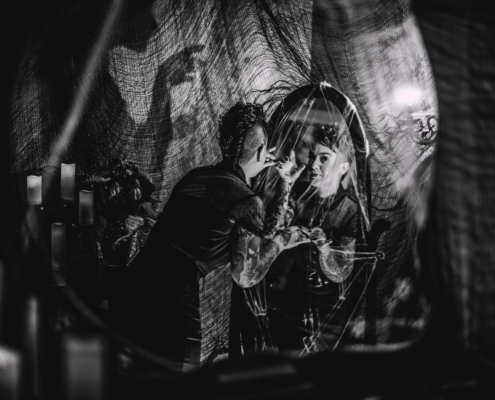 black and white image of a halloween styled room with spider webs, candles, a woman looking in a mirror, and a creepy shadow on the wall beside her