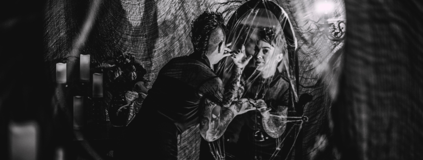 black and white image of a halloween styled room with spider webs, candles, a woman looking in a mirror, and a creepy shadow on the wall beside her