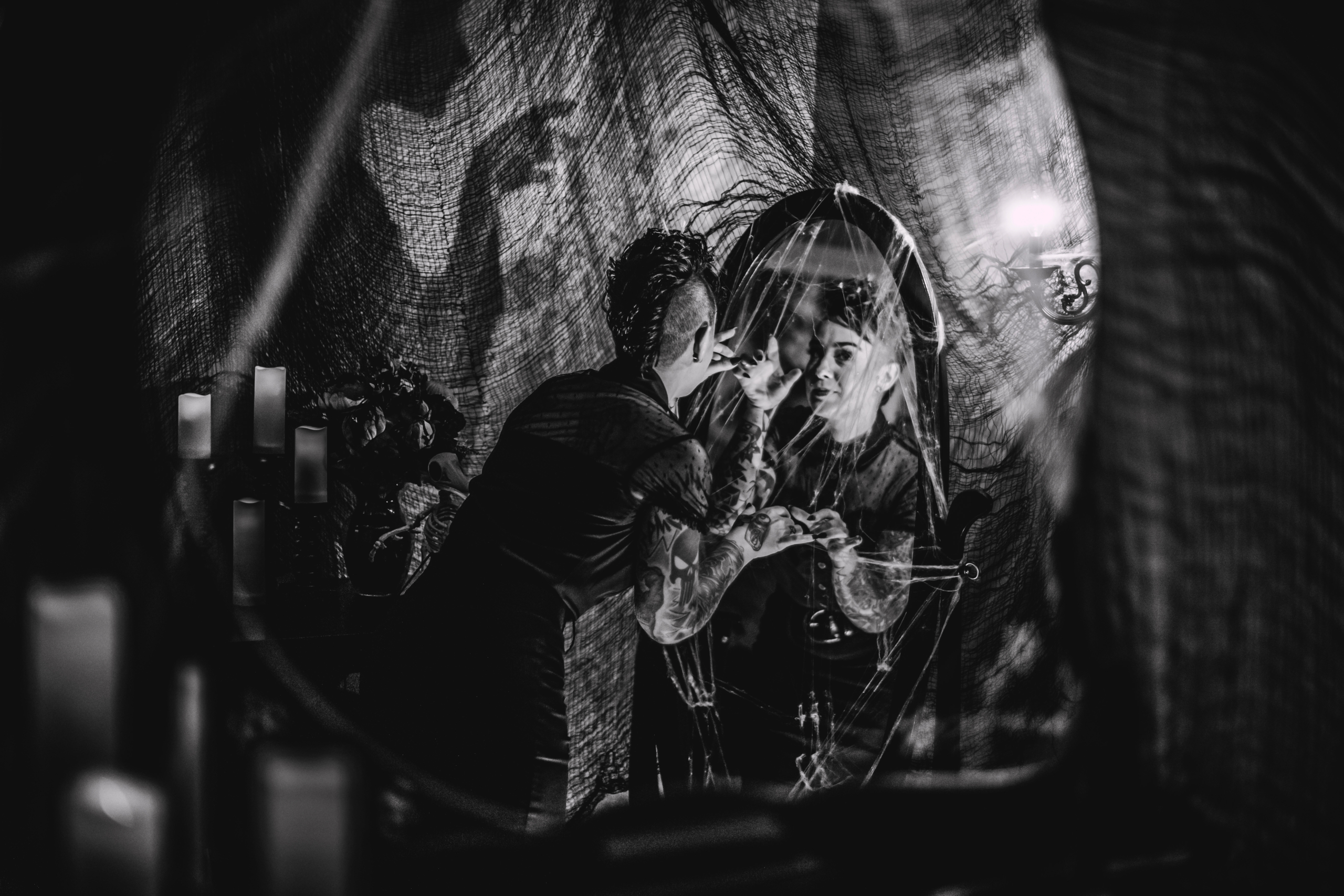 black and white image of a halloween styled room with spider webs, candles, a woman looking in a mirror, and a creepy shadow on the wall beside her