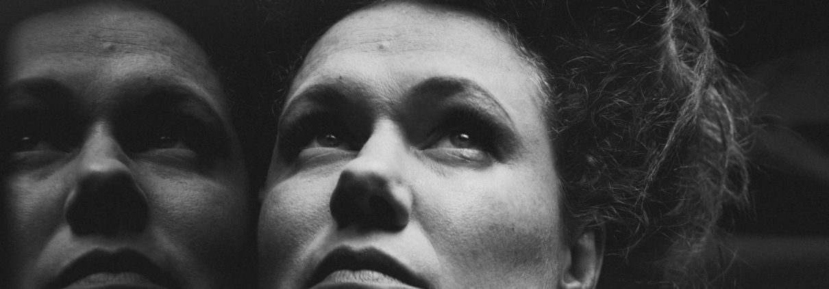 Black and White Close Up of a women's face with a reflection to the left. She has pale skin and freckles and her hair is in a 1910s style piled on top of her head. She is looking up with a pensive and somewhat serious look on her face.