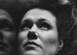 Black and White Close Up of a women's face with a reflection to the left. She has pale skin and freckles and her hair is in a 1910s style piled on top of her head. She is looking up with a pensive and somewhat serious look on her face.