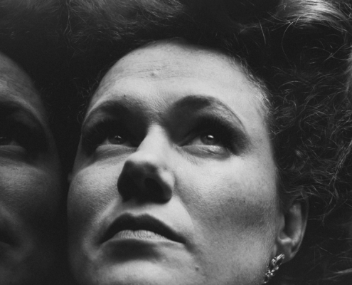 Black and White Close Up of a women's face with a reflection to the left. She has pale skin and freckles and her hair is in a 1910s style piled on top of her head. She is looking up with a pensive and somewhat serious look on her face.