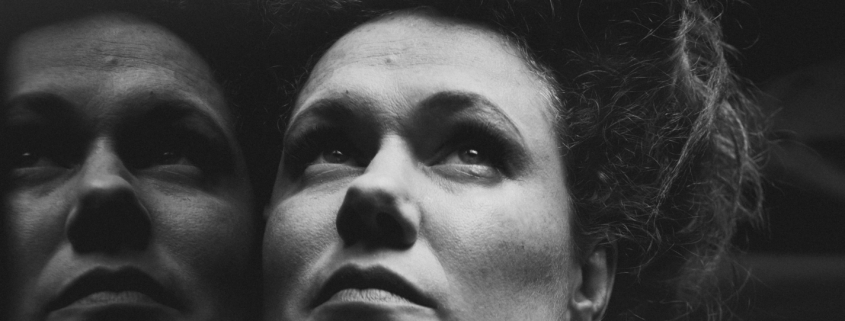 Black and White Close Up of a women's face with a reflection to the left. She has pale skin and freckles and her hair is in a 1910s style piled on top of her head. She is looking up with a pensive and somewhat serious look on her face.