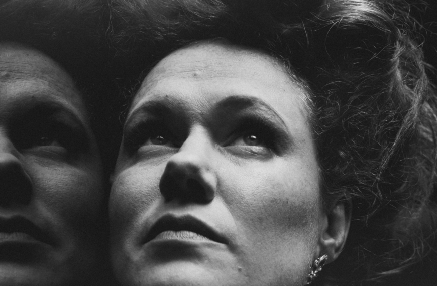 Black and White Close Up of a women's face with a reflection to the left. She has pale skin and freckles and her hair is in a 1910s style piled on top of her head. She is looking up with a pensive and somewhat serious look on her face.